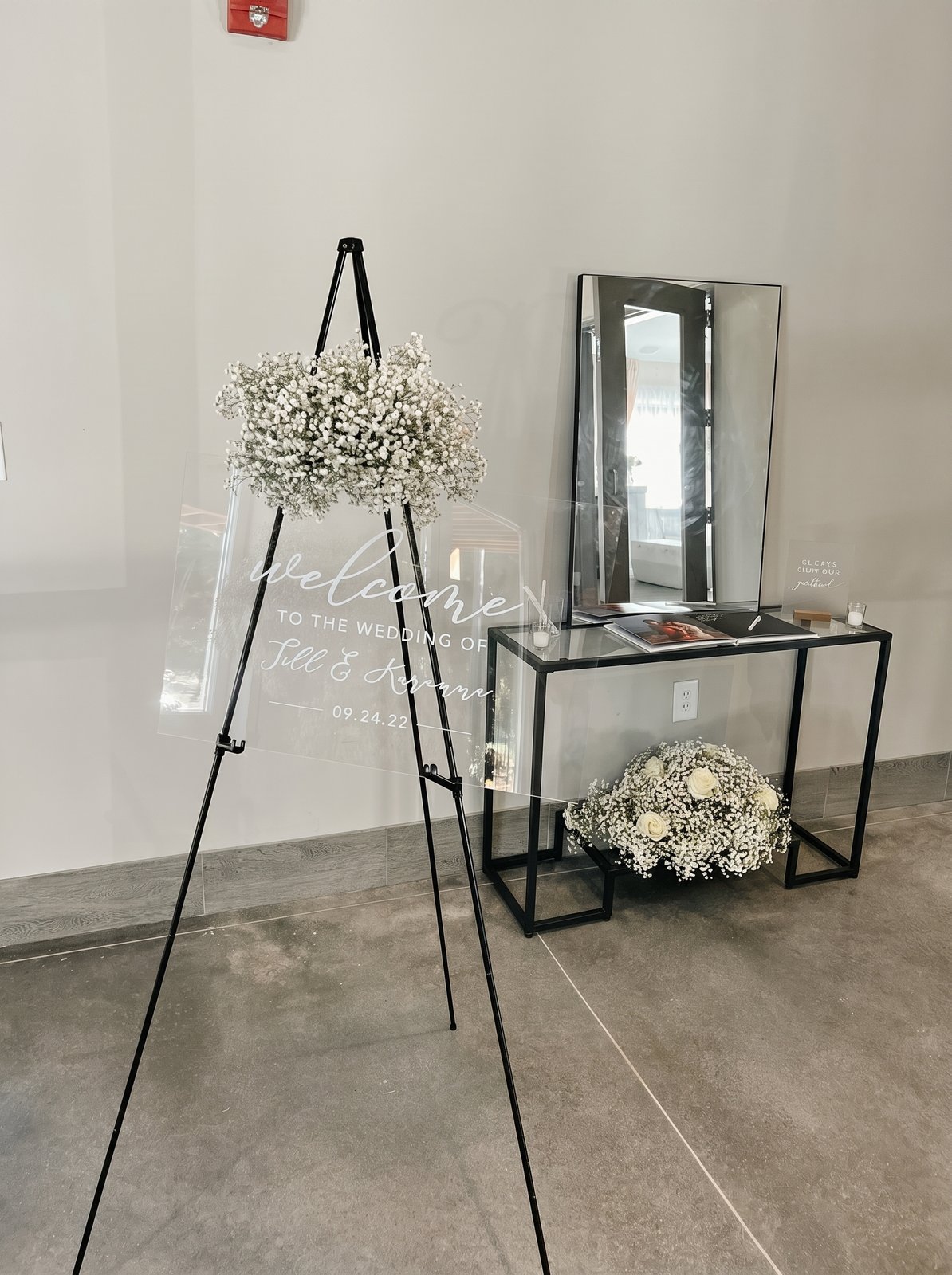 Acrylic wedding welcome sign on an easel beside a gift table with baby's breath and a mirror