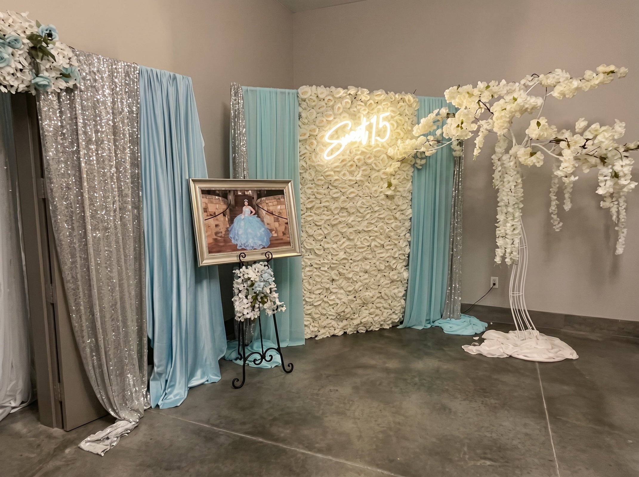 Sweet 15 backdrop with blue draping, a white flower wall, neon sign and a portrait on an easel