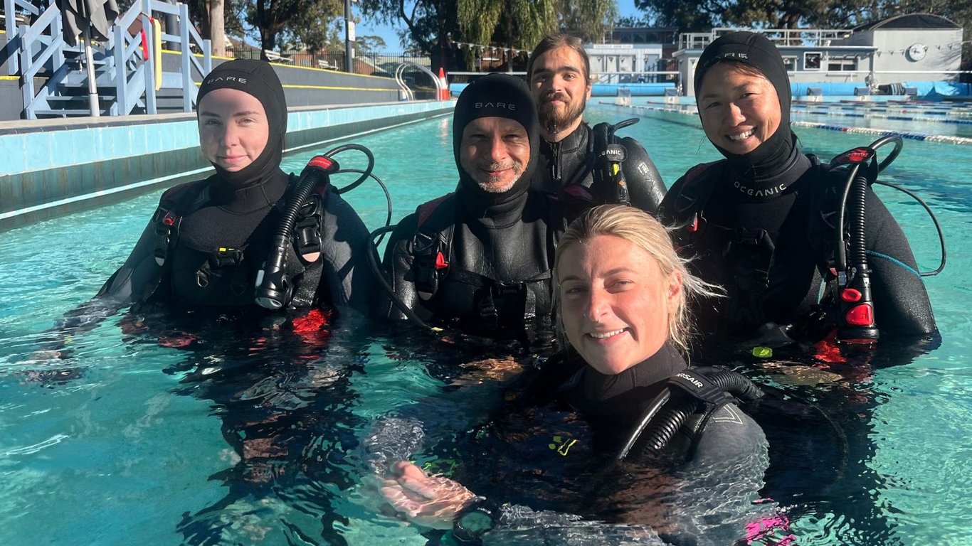 A scuba diving instructor is demonstrating the proper mask clearing technique to a small group of students in a swimming pool, surrounded by scuba diving equipment. This fun experience is part of their training in a dive course, helping complete beginners learn essential skills for their scuba diving adventures.