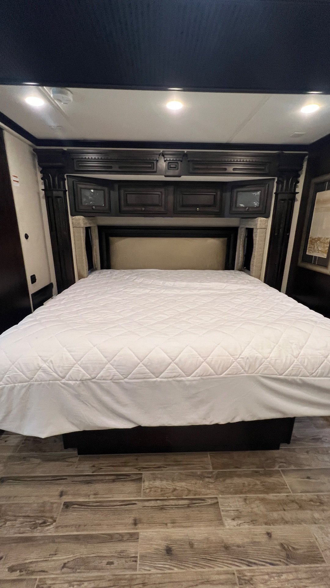 2018 Newmar Dutch Star 4369 — Main bedroom with a large made bed, upholstered headboard, overhead cabinets, and wood-look flooring