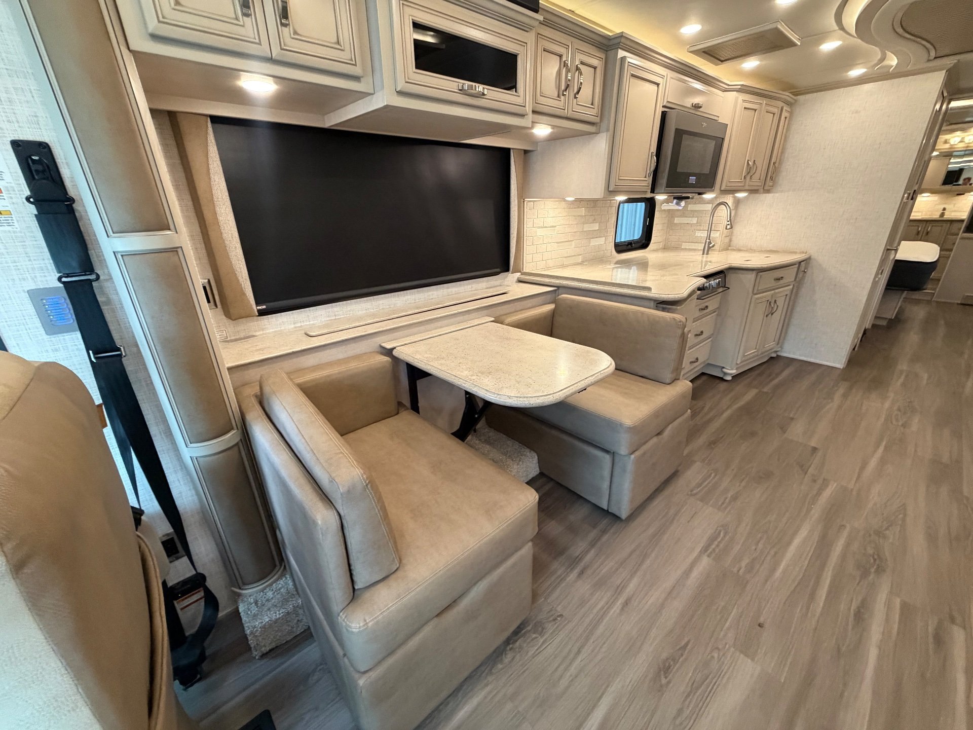 2023 Newmar Kountry Star 4068 — Dinette beside a large television with adjacent kitchen counter, sink, microwave, and cabinetry