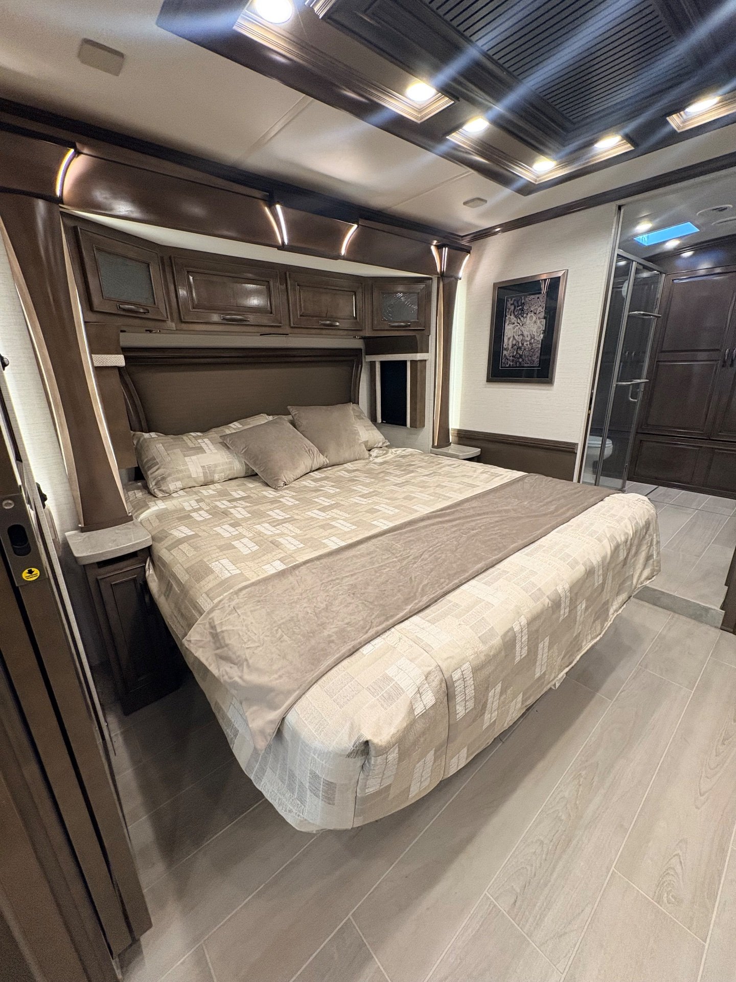 2025 Newmar Dutch Star 4369 — Main bedroom with a made bed, overhead cabinets, bedside storage, and an adjoining bathroom doorway