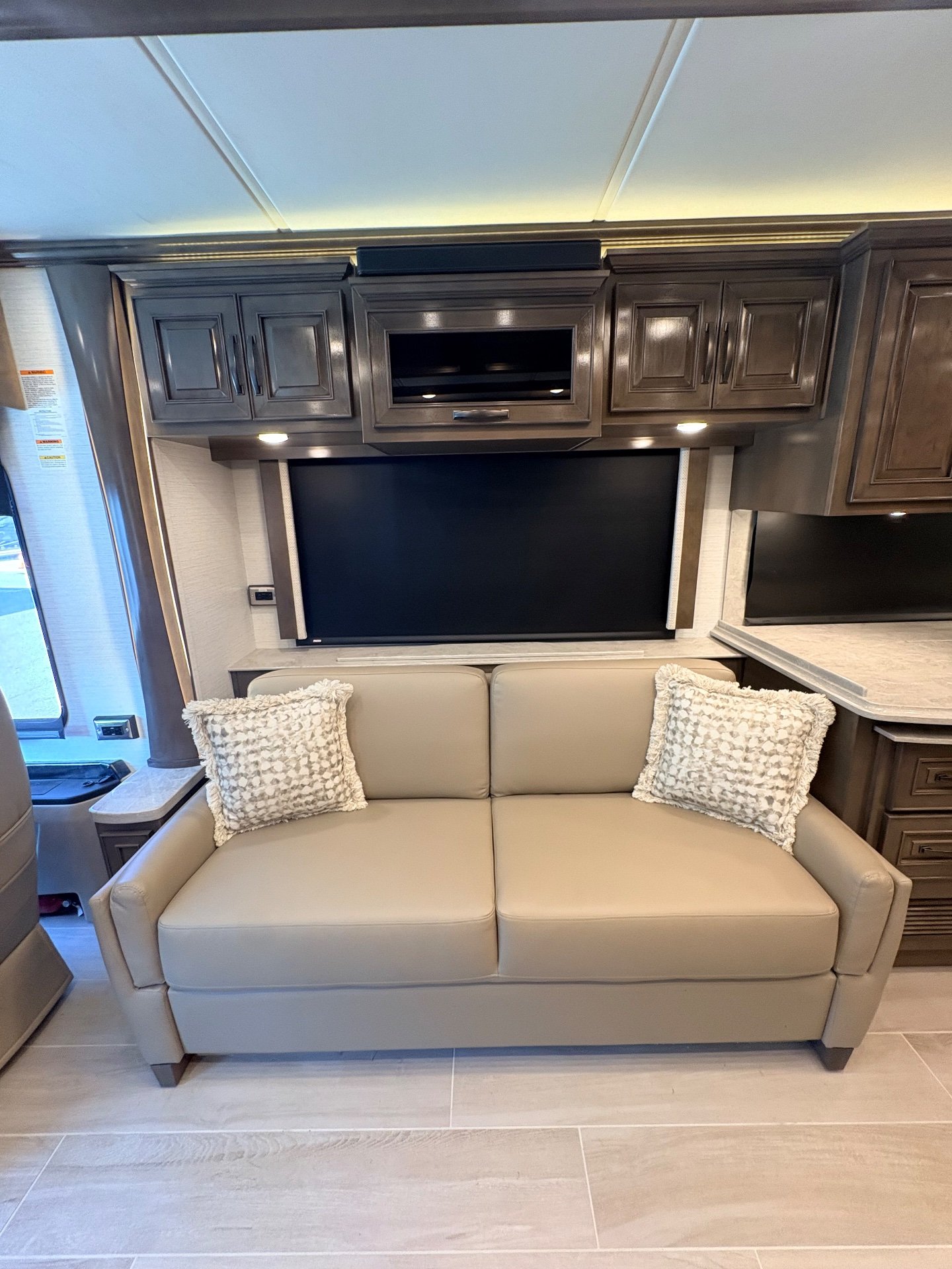 2025 Newmar Dutch Star 4369 — Lounge sofa beneath a large television, upper cabinets, and a covered window beside the kitchen counter