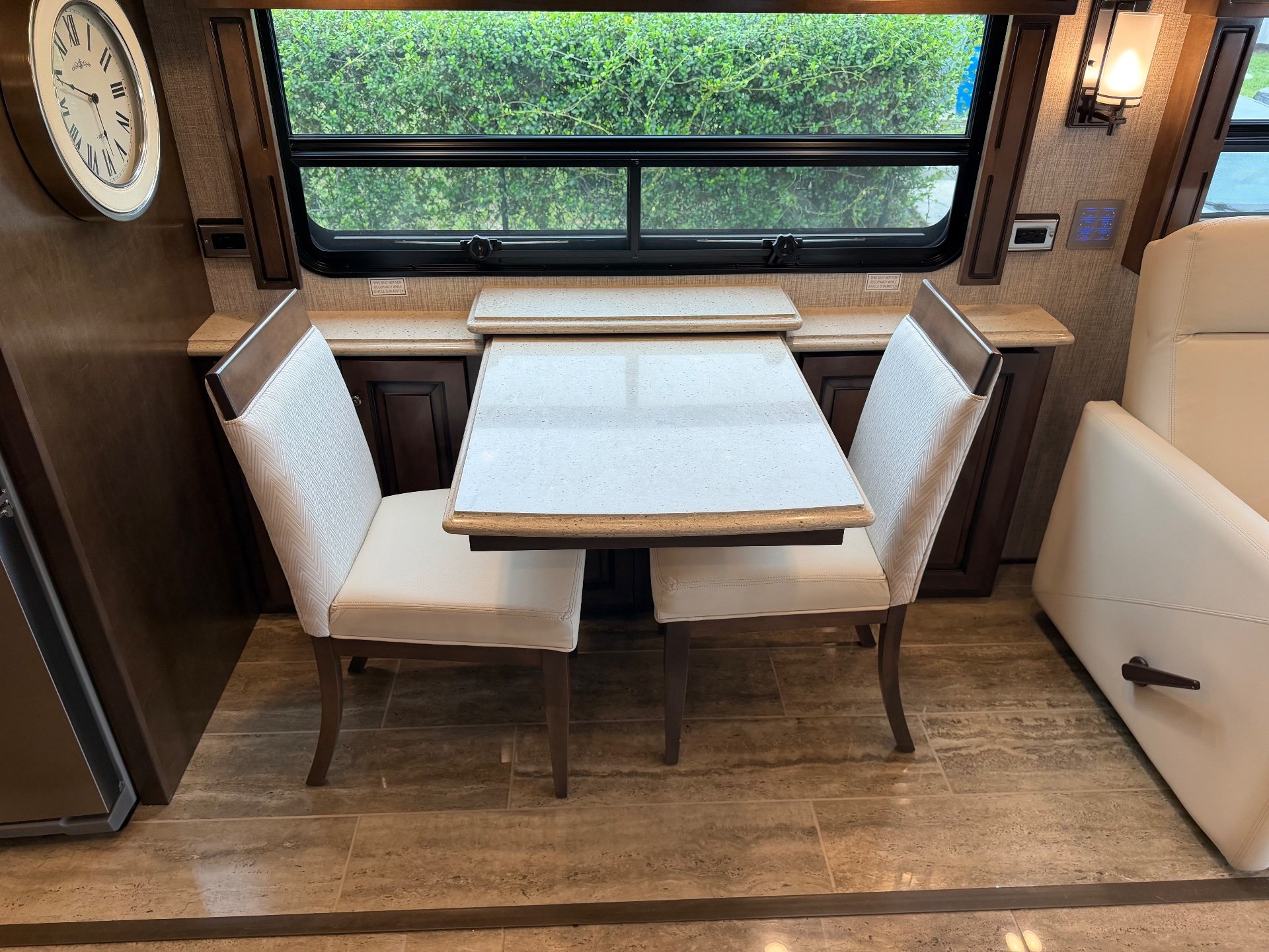2021 Newmar Dutch Star 4369 — Freestanding dining table and two chairs beside a cream lounge recliner under a large window