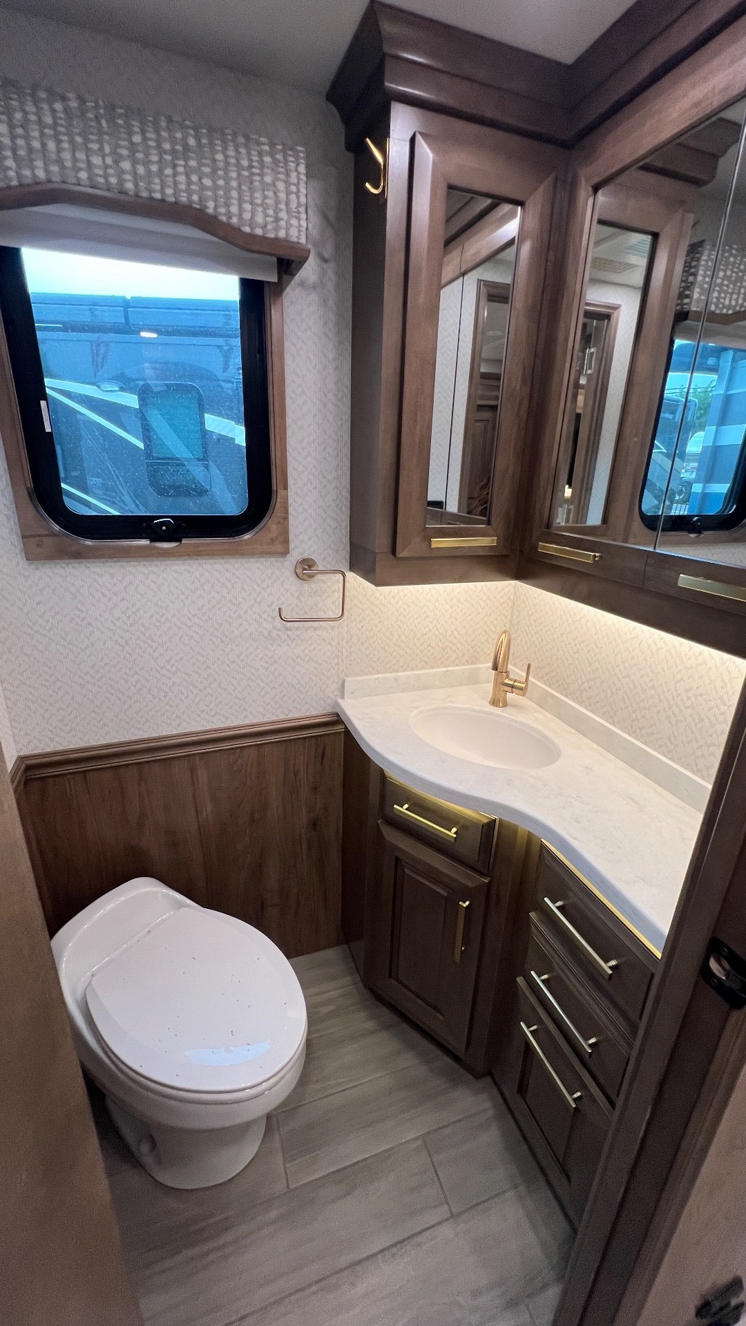 2023 Entegra Coach Cornerstone 45B — Compact bathroom with toilet, vanity sink, mirrored cabinets, window, and illuminated countertop