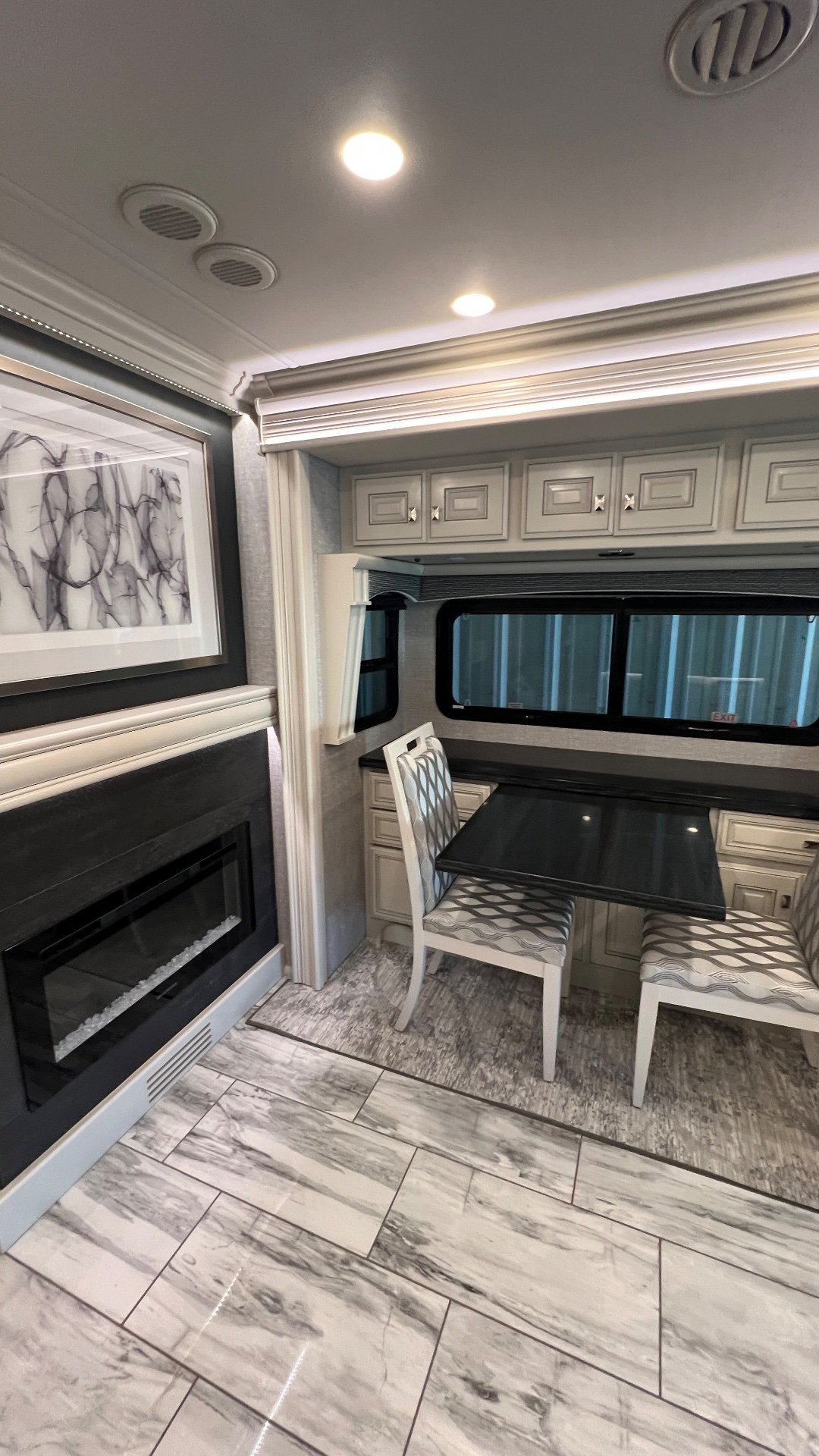 2024 Fleetwood Discovery LXE 40M — Dining table and two chairs beside a fireplace, with overhead cabinets and lounge flooring visible