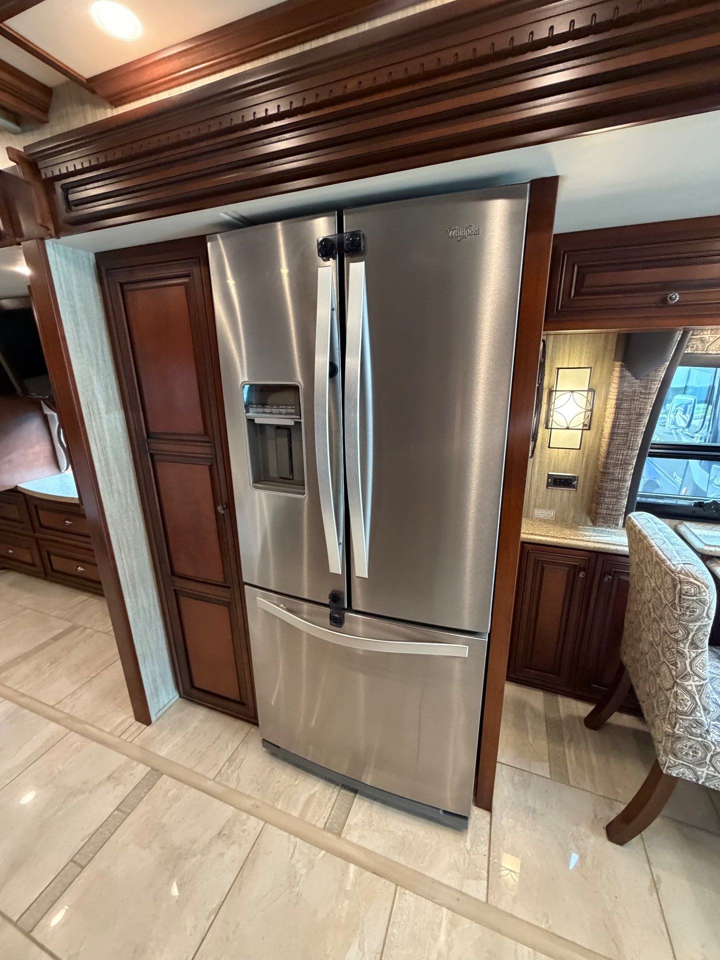 2017 Newmar Dutch Star 4369 — Stainless refrigerator beside pantry cabinetry, with a dining chair, window, and dinette cabinetry visible nearby