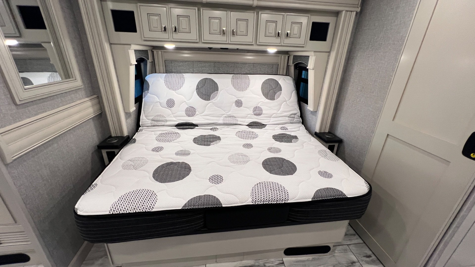 2024 Fleetwood Discovery LXE 40M — Main bedroom with a patterned mattress, bedside tables, overhead cabinets, and a wardrobe door