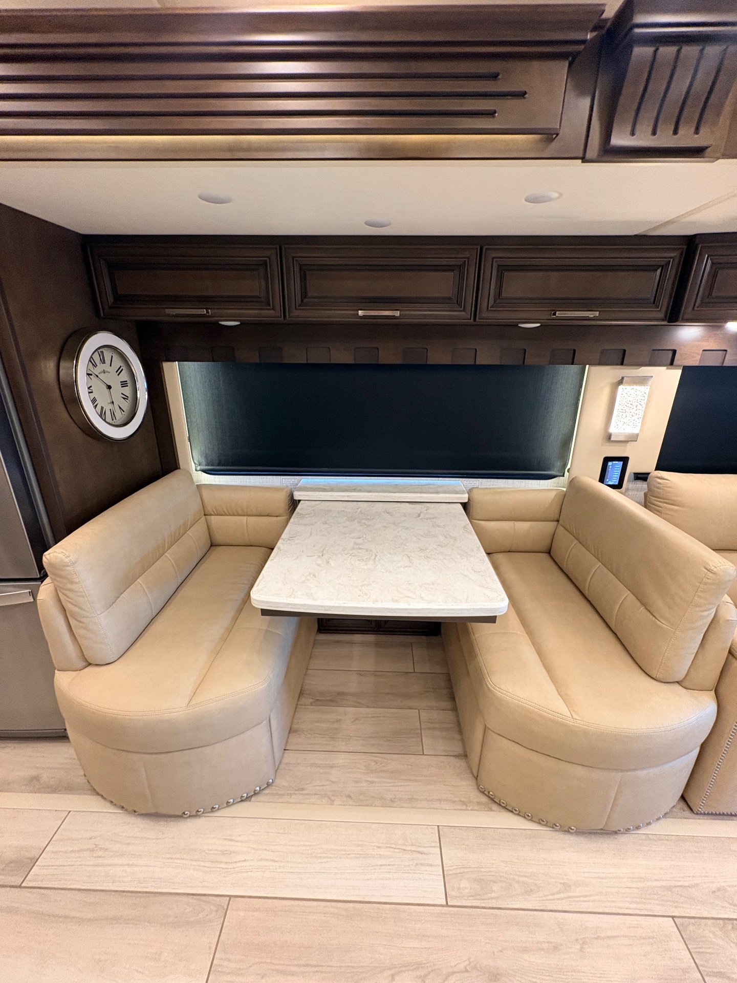 2023 Newmar London Aire 4579 — Motorhome dinette with opposing upholstered benches, table, window shade, wall clock, and overhead cabinetry