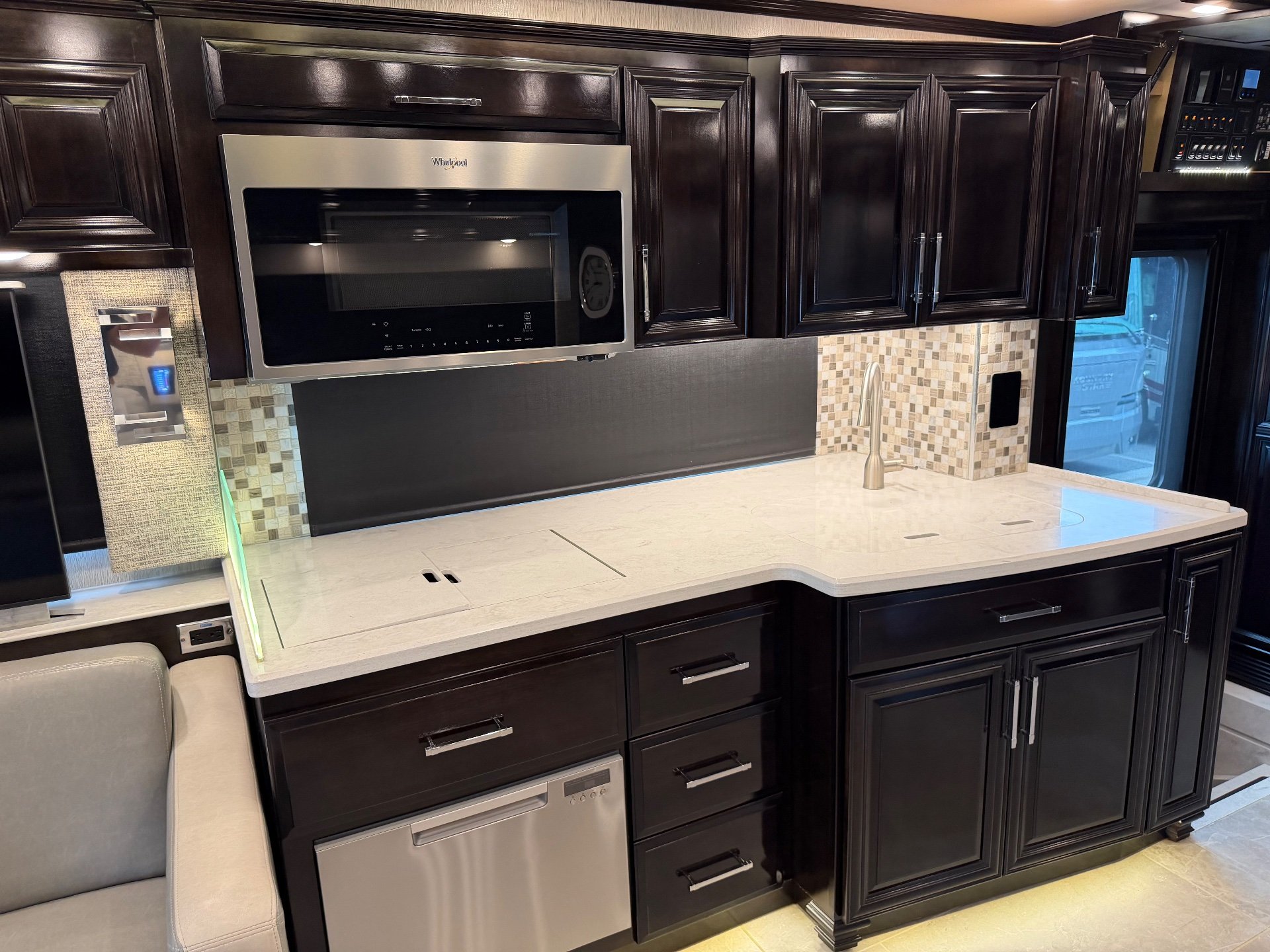 2023 Newmar Supreme Aire 4575 — Motorhome kitchen with broad countertop, sink, microwave, dishwasher, mosaic backsplash, drawers, and cabinetry