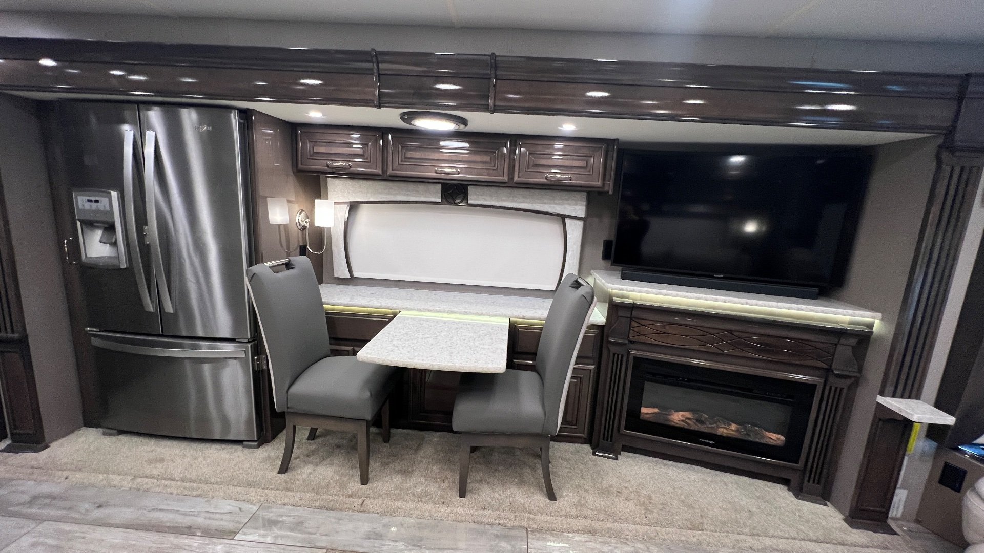 2021 Entegra Coach Cornerstone 45B — Dining table and chairs between a refrigerator and fireplace television wall in the motorhome lounge