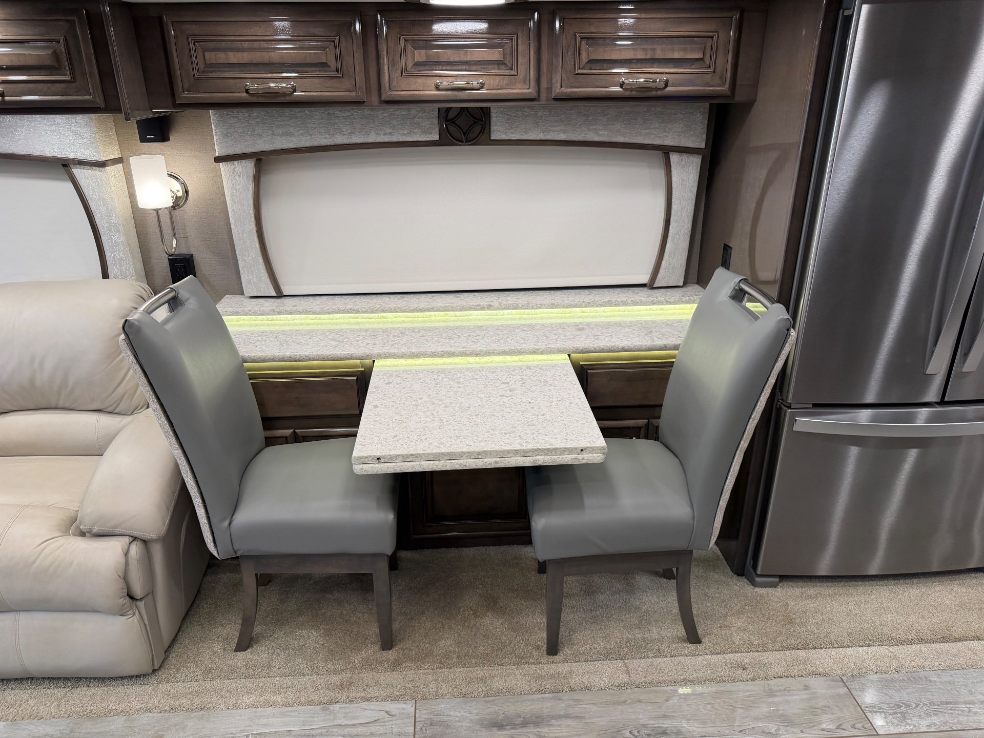2021 Entegra Coach Cornerstone 45F — Dining table and two chairs beside a recliner and full-height refrigerator in the main living area