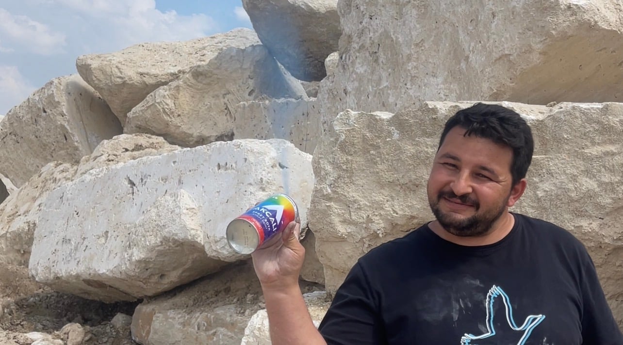 A man in a black T-shirt standing among rough quarried stone blocks, raising a drink can towards the camera.
