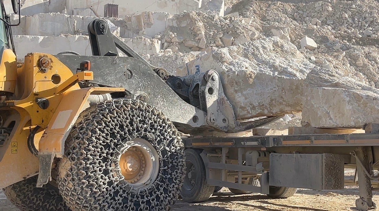 A wheel loader on chained tyres lowering a rough stone block onto a flatbed trailer at a quarry.