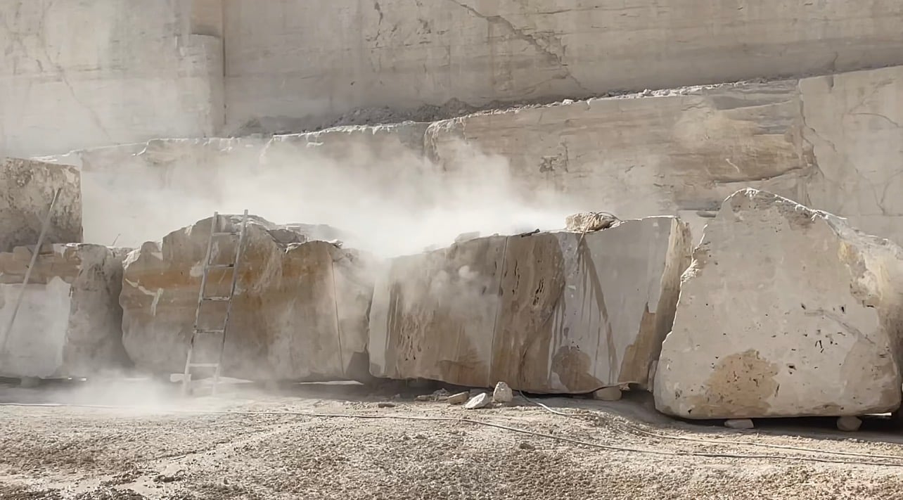 Large pale stone blocks lined up at the foot of a quarry face, with a haze of stone dust drifting between them.