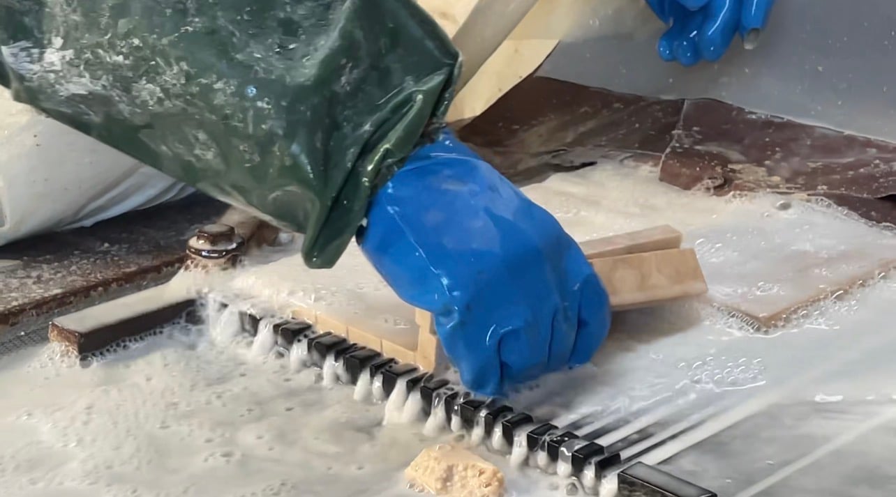 A gloved hand guiding a strip of stone along a wet saw, water foaming over the blade and the freshly cut pieces.