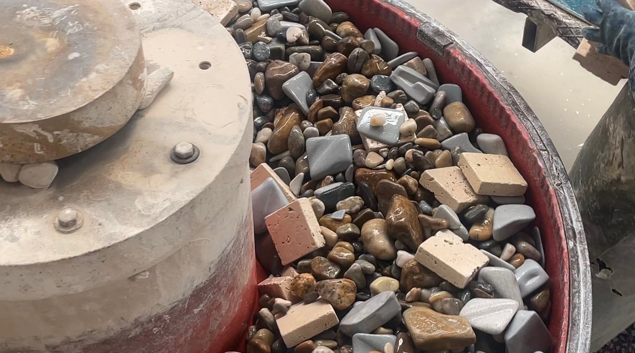 A tumbling drum holding small square cut stone tiles among smooth grey and brown pebbles.