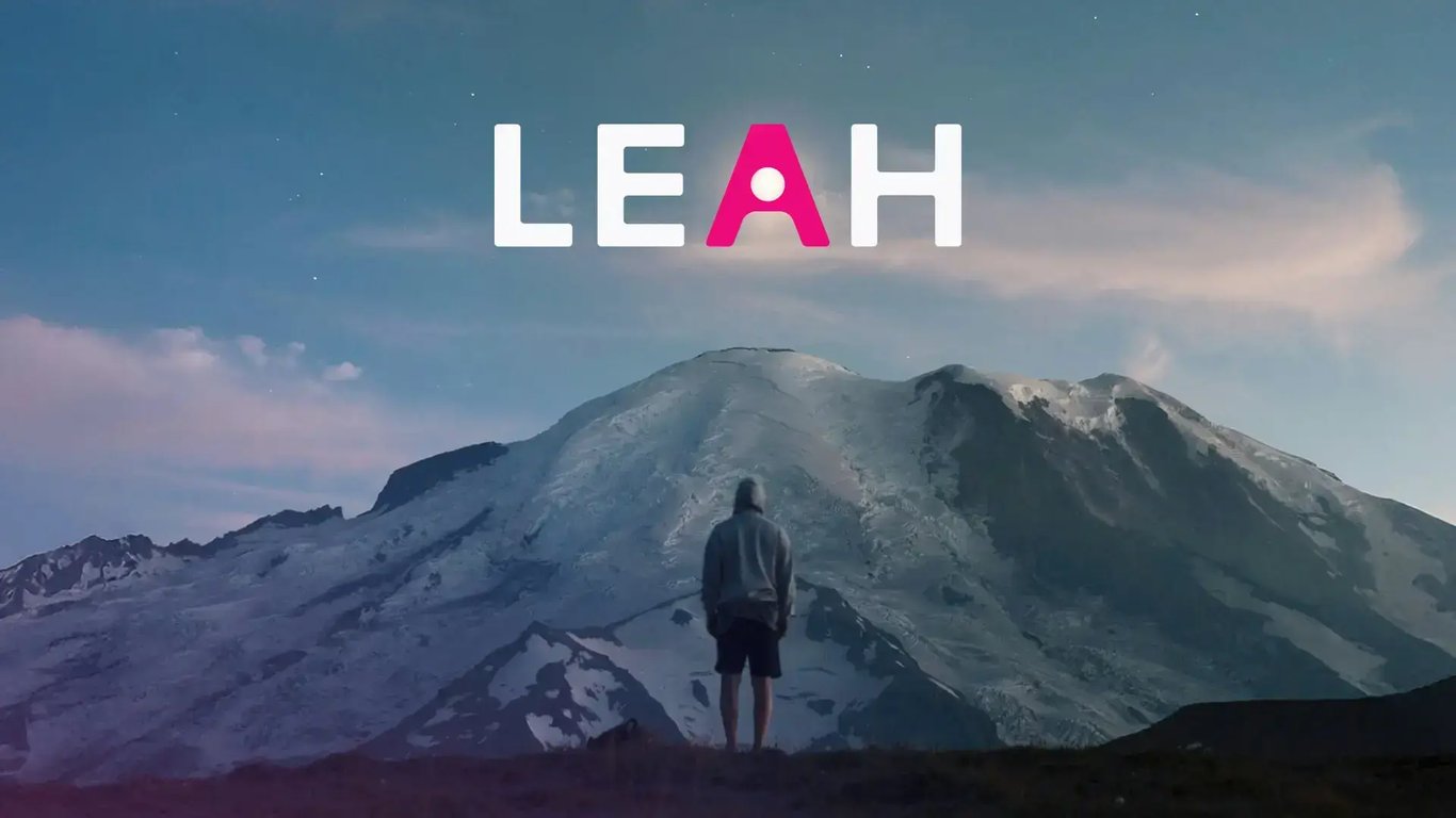 Person in a hoodie stands before a snow-covered mountain at dusk, with the word “LEAH” in bold white and pink letters above the peak.