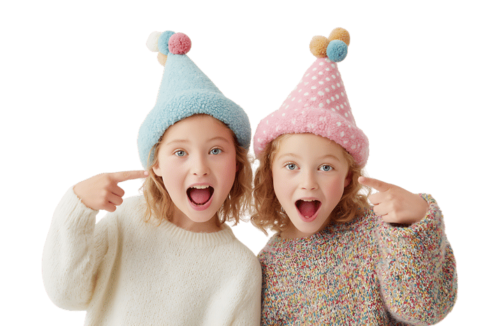 Two girls in party hats point at themselves.