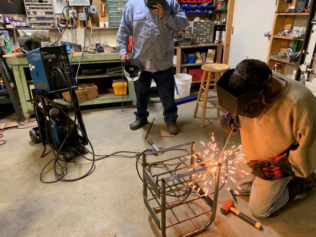 Jimmie stick-welding on a metal rack in the shop, while his first client looks on