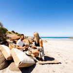 Couple enjoying a cozy picnic on Kangaroo Island's beach. They sit together on a blanket surrounded by a spread of fresh, local treats, with the ocean in the background. A beautiful, relaxed setting for a romantic moment amidst the island's scenic views and natural surroundings