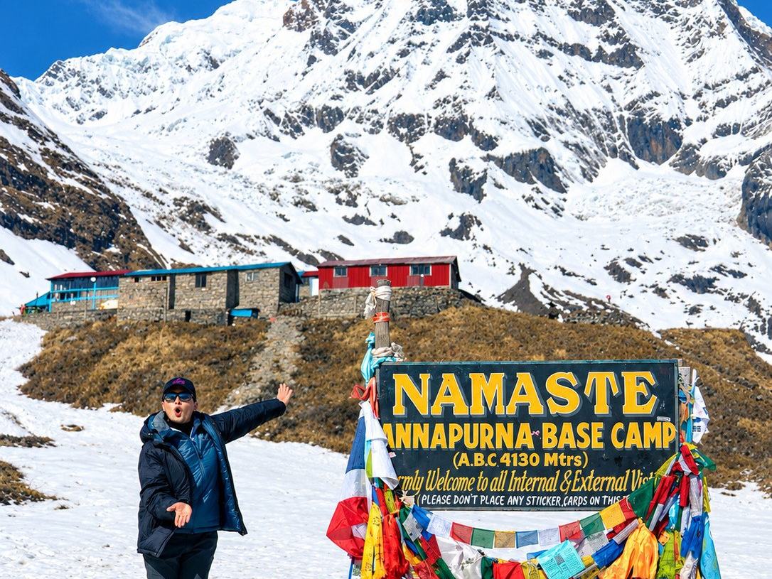 A trekker with an arm raised beside the Namaste Annapurna Base Camp sign and its prayer flags, the snow face of Annapurna South filling the sky above the camp huts