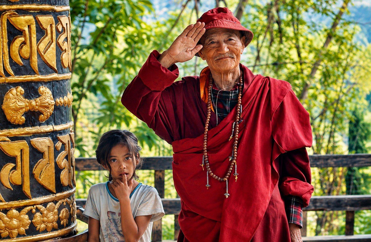 An elderly man in a red robe saluting beside a prayer wheel, with a child, Bhutan