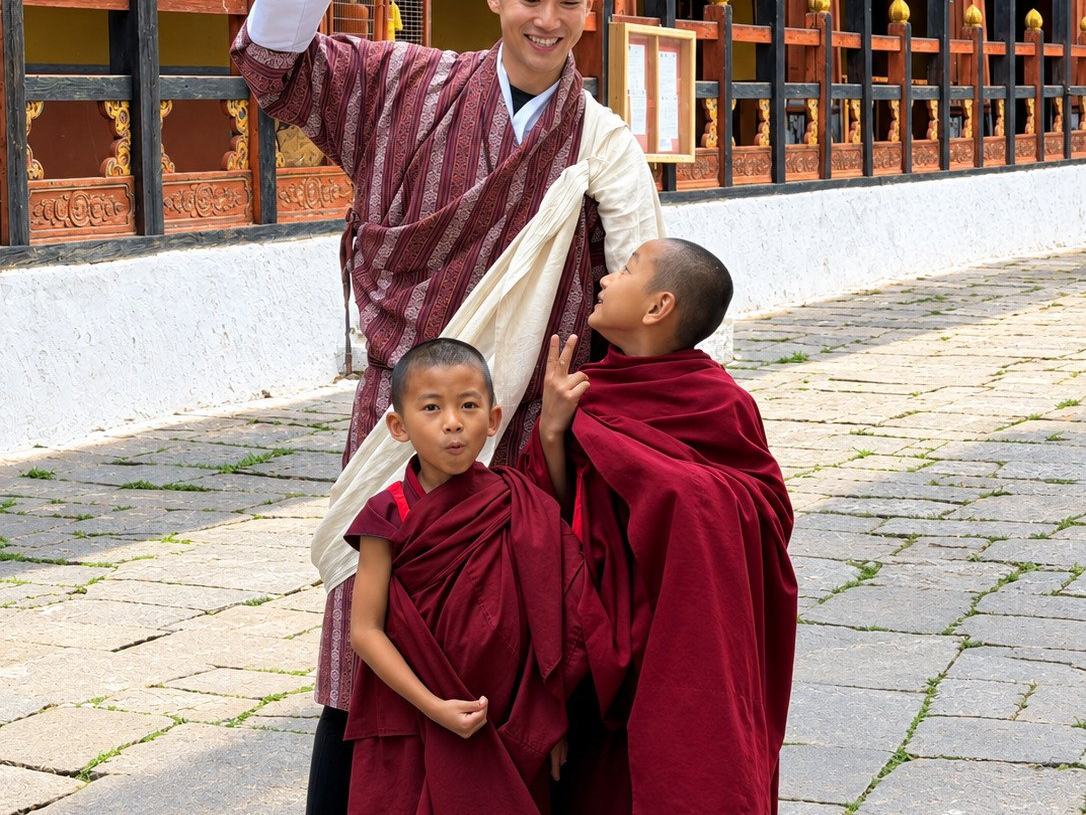 A Bhutanese guide in a gho pointing something out to two young novice monks in red robes, in the courtyard of Rinpung Dzong in Paro