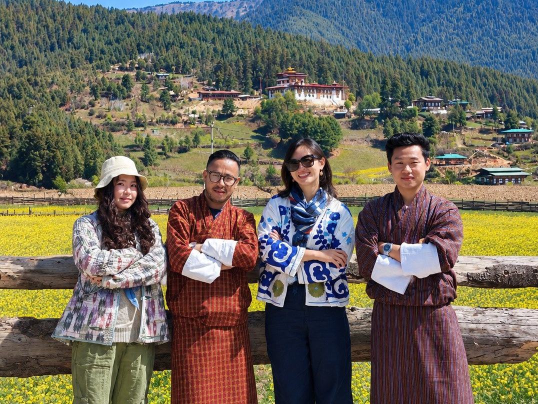 Two travellers standing with their Bhutanese hosts in gho at a timber fence above a field of mustard flowers, a hillside monastery and forested ridges rising behind them