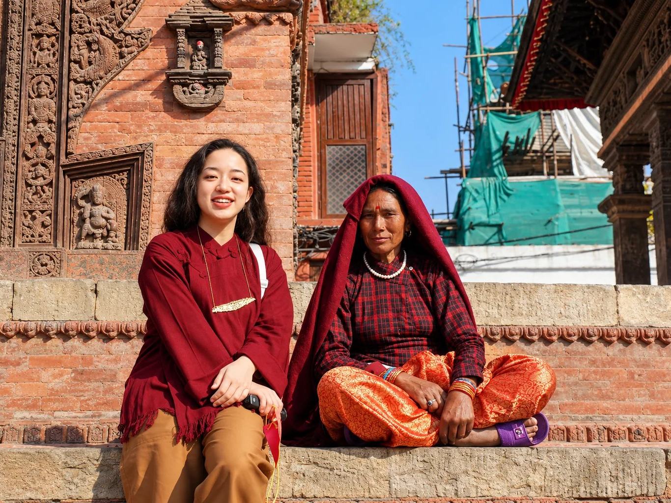 A traveller and a local woman seated on carved temple steps in a Kathmandu Valley Durbar Square, Nepal