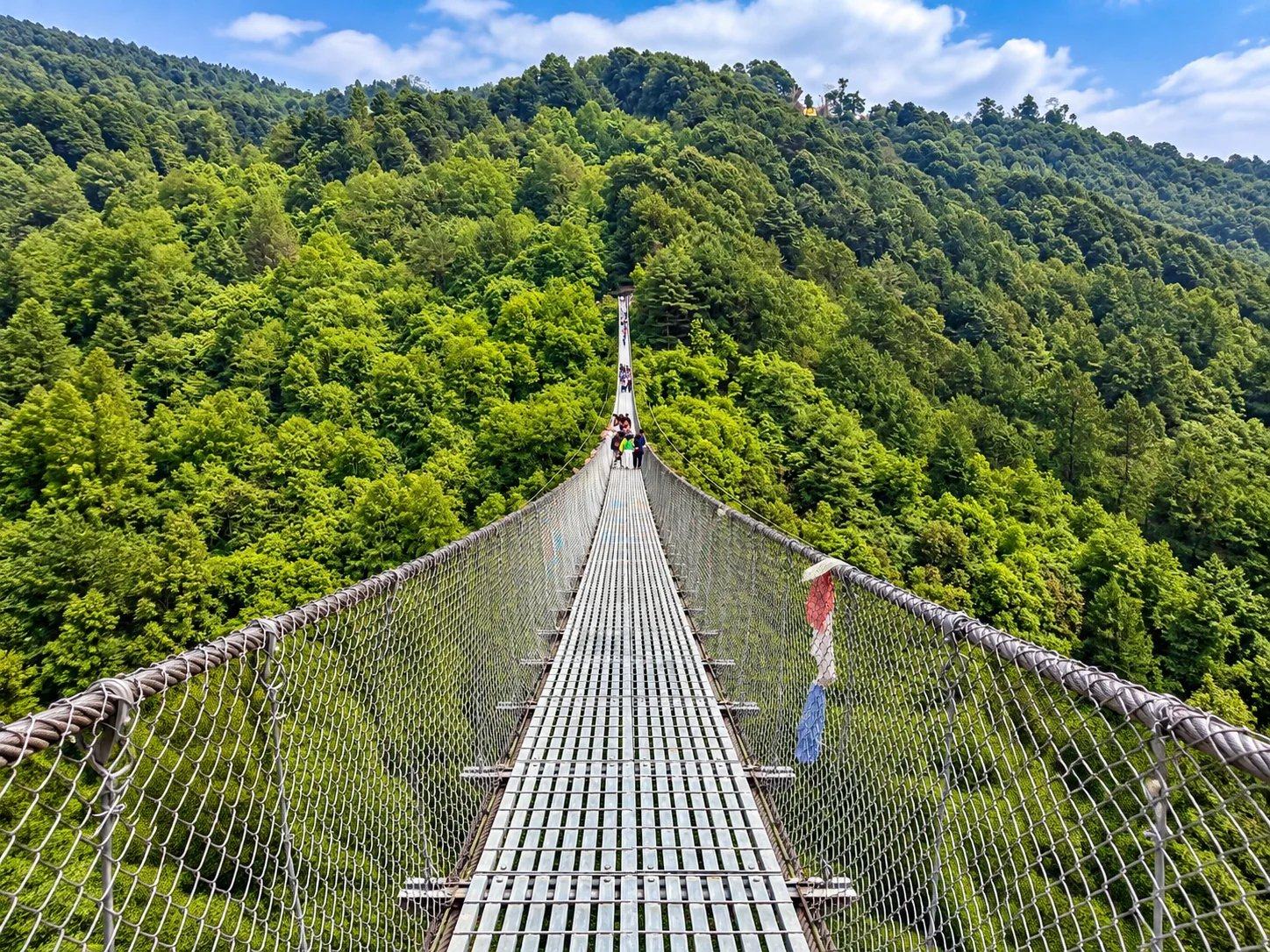 A steel suspension bridge stretching across a forested valley in the hills of Nepal