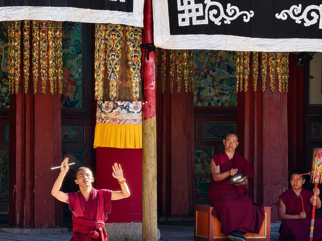 A monk in maroon robes mid cham dance in a monastery courtyard in Tibet, musicians seated beneath painted timber pillars and black appliqué banners