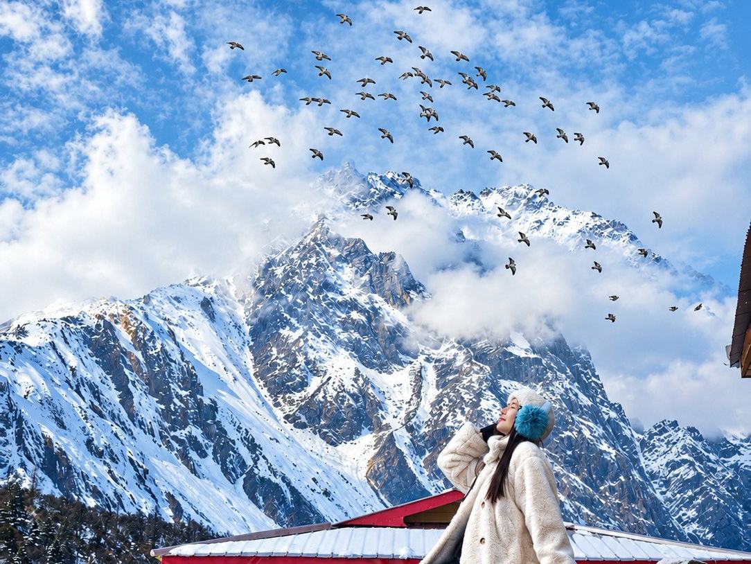 A traveller resting on a stone wall beside a painted temple roof, a snow-covered Himalayan face and a flock of birds filling the sky behind