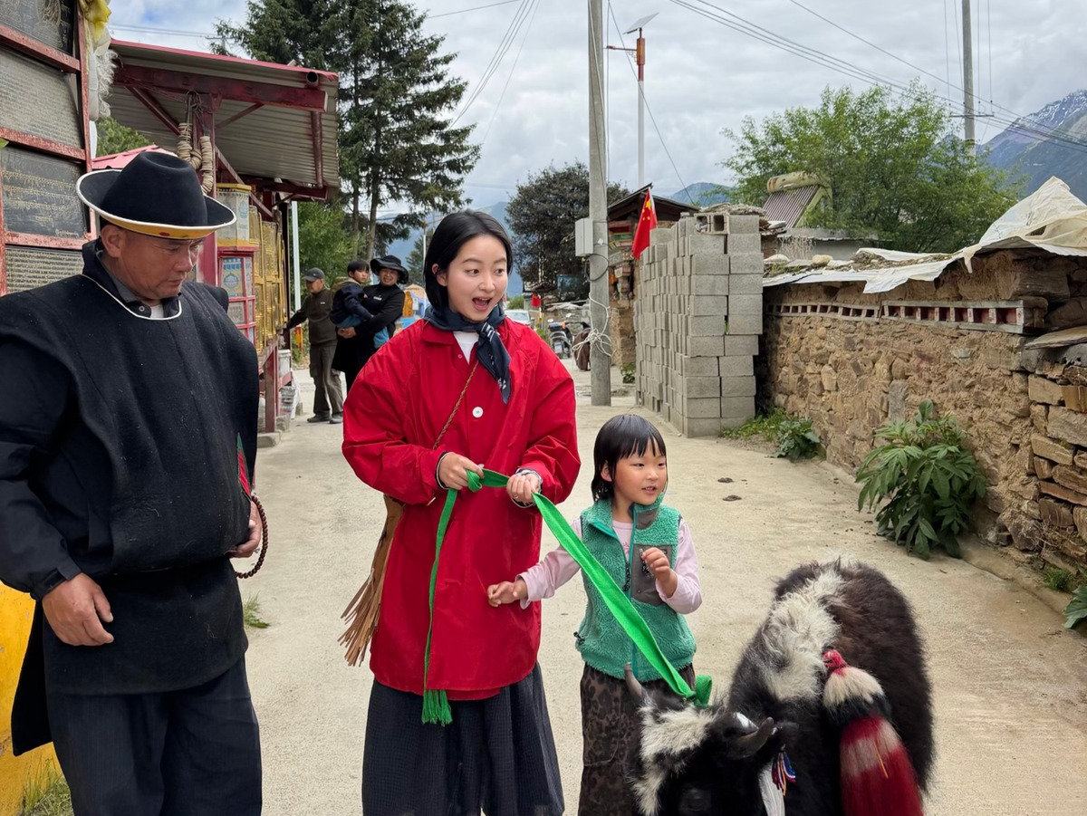 A traveller in a red jacket meeting a Tibetan elder and a child leading a decorated yak along a village lane on the plateau
