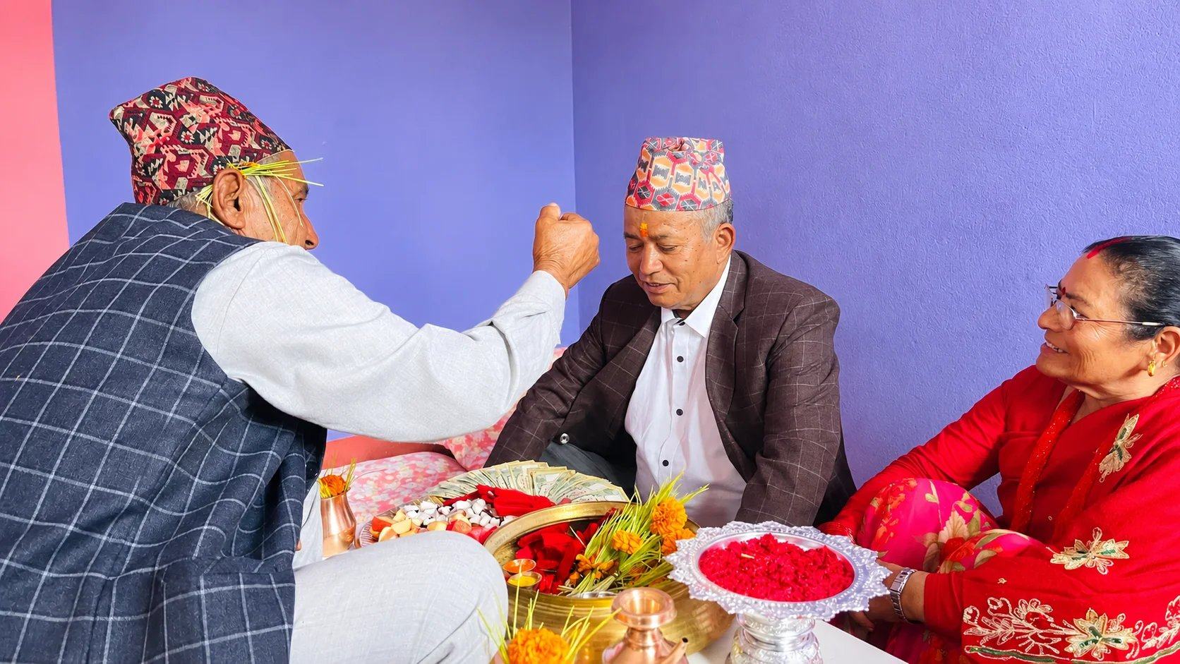 An elder with jamara behind his ear gives a Dashain blessing to a man in a dhaka topi, beside a tray of red tika and marigolds, Nepal
