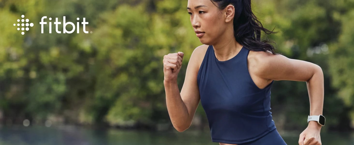 A woman running outdoors near water, wearing a navy athletic crop top and the Fitbit Sense 2 Advanced Health Smartwatch in white. The Fitbit logo appears in the upper left corner against a blurred green background.