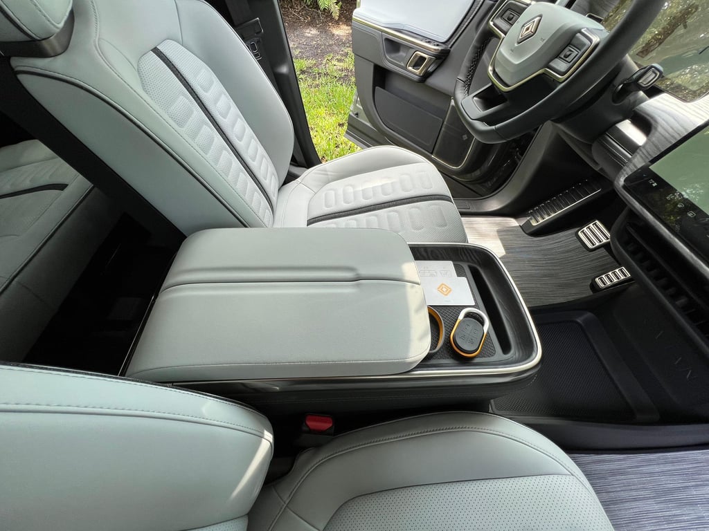 Rivian R1S front interior close-up showing light gray perforated seats, center console storage, premium trim, and the driver cockpit area with steering wheel and pedals visible.