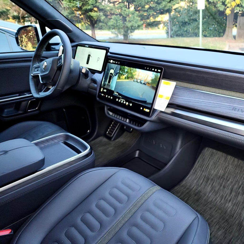 Modern Rivian R1S interior featuring a minimalist dashboard with dual digital displays, large central touchscreen showing camera view, steering wheel controls, premium seats, and a clean, tech-focused cabin design.