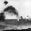 Black-and-white photograph of a naval battle scene with thick smoke, a low-flying aircraft, and water plumes from near-misses around a ship under attack at sea