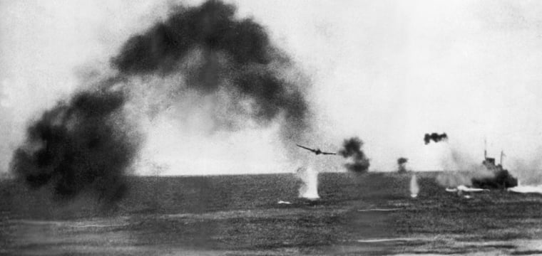 Black-and-white photograph of a naval battle scene with thick smoke, a low-flying aircraft, and water plumes from near-misses around a ship under attack at sea