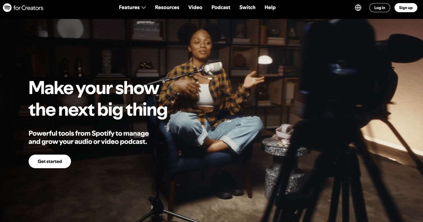 A person sits in a blue chair, recording a podcast with a microphone, surrounded by shelves and a camera focused on them.