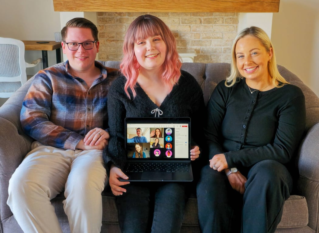 Three members of the Inkfire team sitting together on a couch, holding a laptop that shows the rest of the team joining by video call.