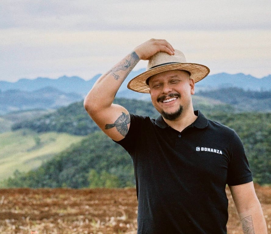 Talles Carvalho of Fazenda Bonanza smiling in a Bonanza-branded polo shirt, standing in front of the hills of Minas Gerais, Brazil.