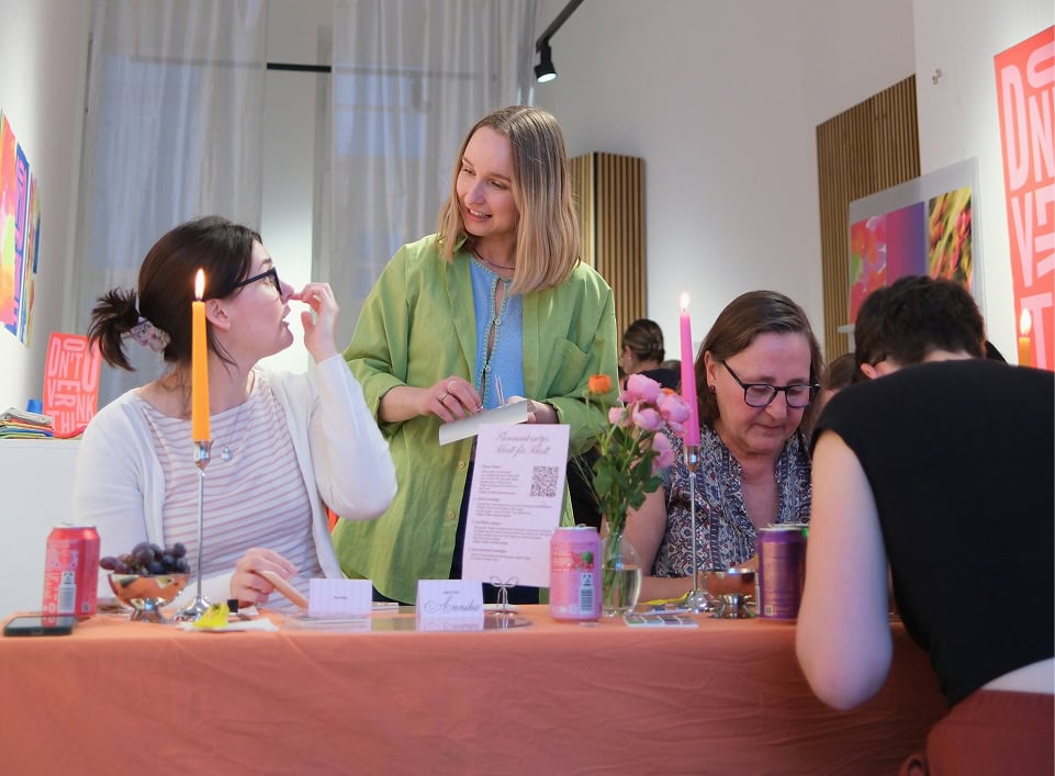 A MuseDuo in-person craft event, with a host in a green jacket helping two women at a candlelit table set with drinks, flowers, and DIY materials.