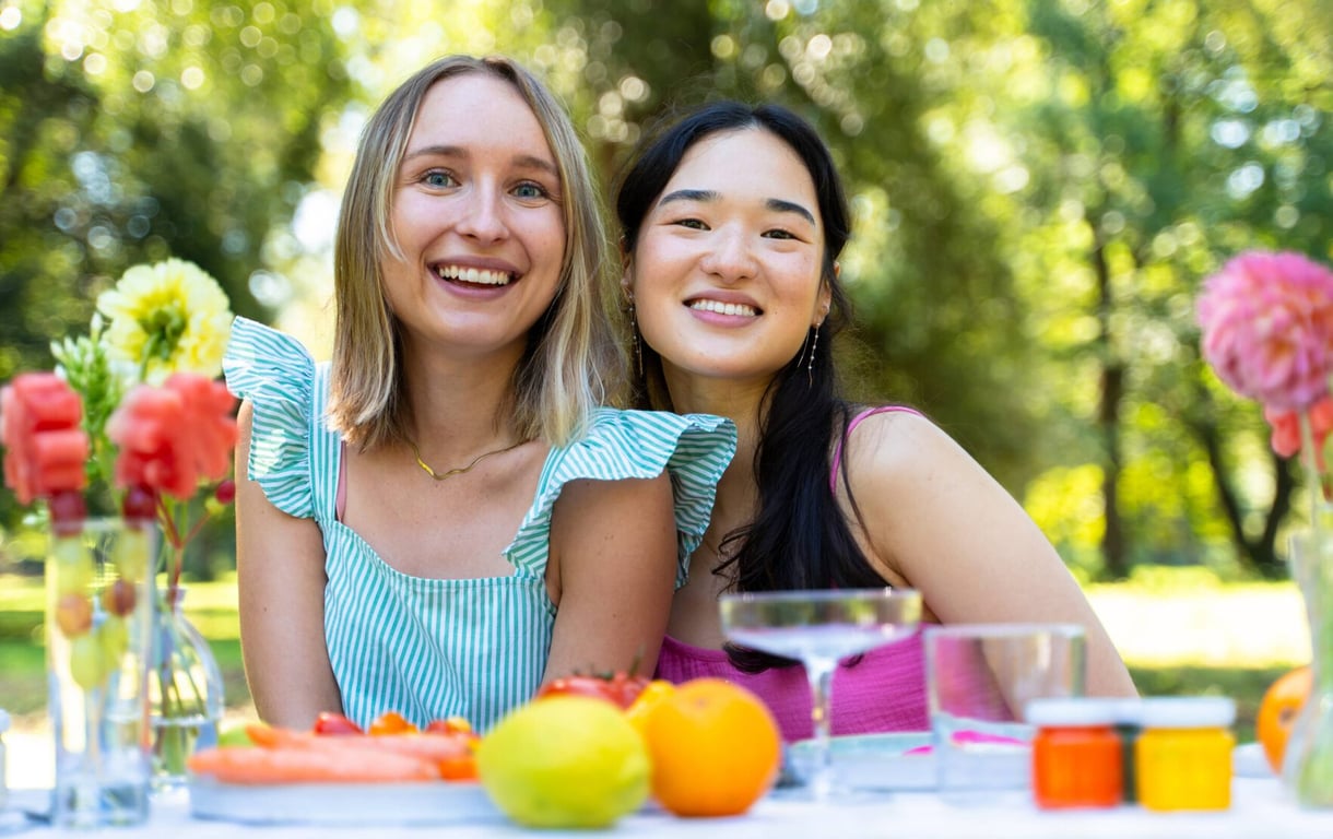 MuseDuo founders Hannah and Nina smiling at an outdoor table set with fruit, flowers, and drinks.
