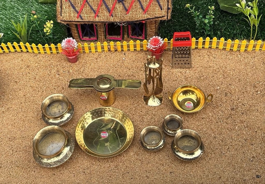 A set of miniature brass cooking pieces, including plates, bowls, a kadai, and a grater, arranged on sand in front of a small decorated toy house, each stamped with the Shripad brand label.