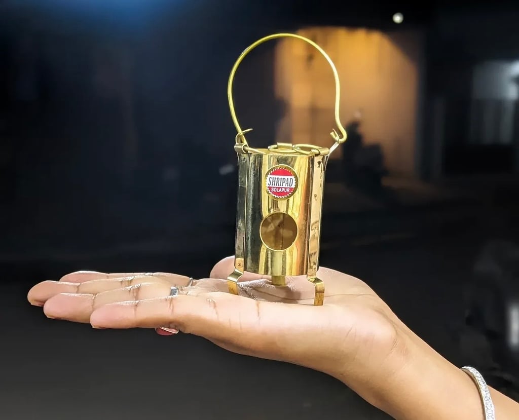 A hand holds a small brass miniature lantern with a carrying handle, branded with a red "Shripad Solapur" label, showing how tiny the piece is against an open palm.