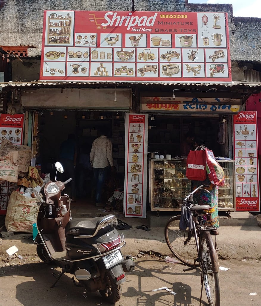 The storefront of Shripad Steel Home in Solapur, with a large red sign reading "Shripad Steel Home, Miniature Brass Toys" above display cases of brass items, a parked scooter and bicycle out front, and a customer browsing.