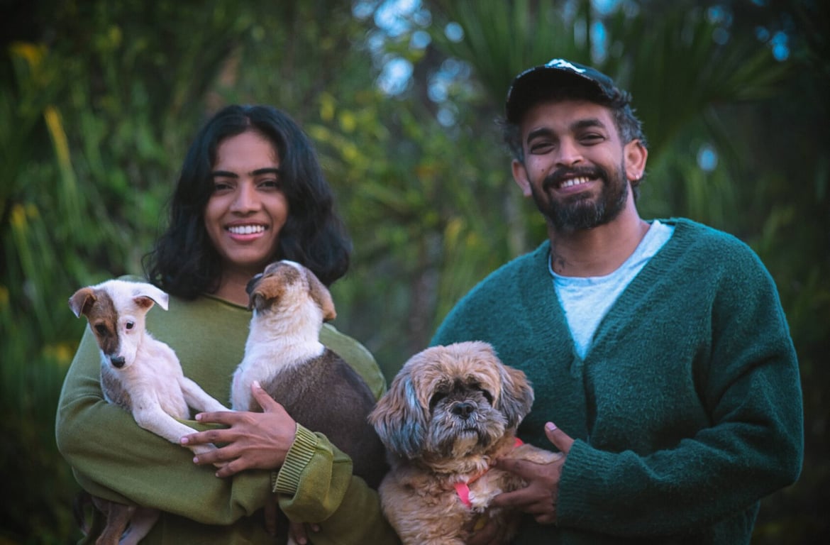 Wilding founders Sree and Sachin smiling outdoors, each holding dogs, with green foliage behind them.