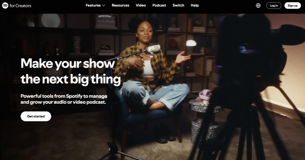Une personne est assise sur une chaise bleue et enregistre un podcast avec un microphone, entour&eacute;e d&rsquo;&eacute;tag&egrave;res et film&eacute;e par une cam&eacute;ra point&eacute;e sur elle.